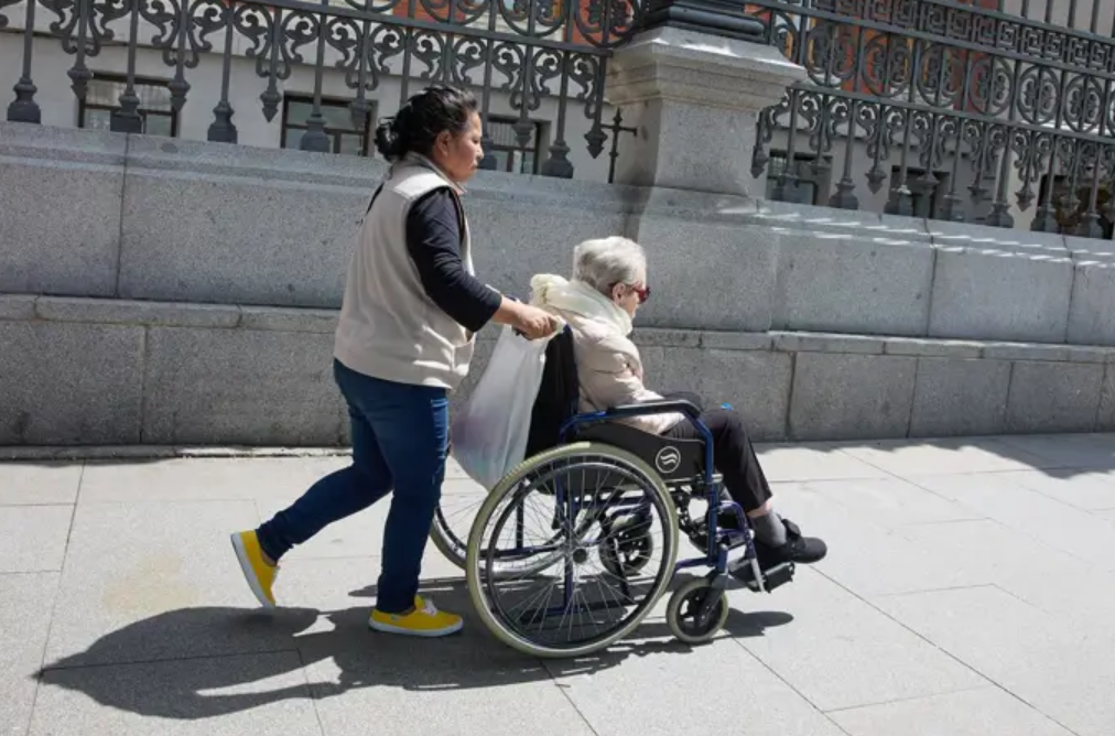 Imagen de una mujer mayor en silla de ruedas junto a su cuidadora paseando un 4 de junio de 2025 en Madrid