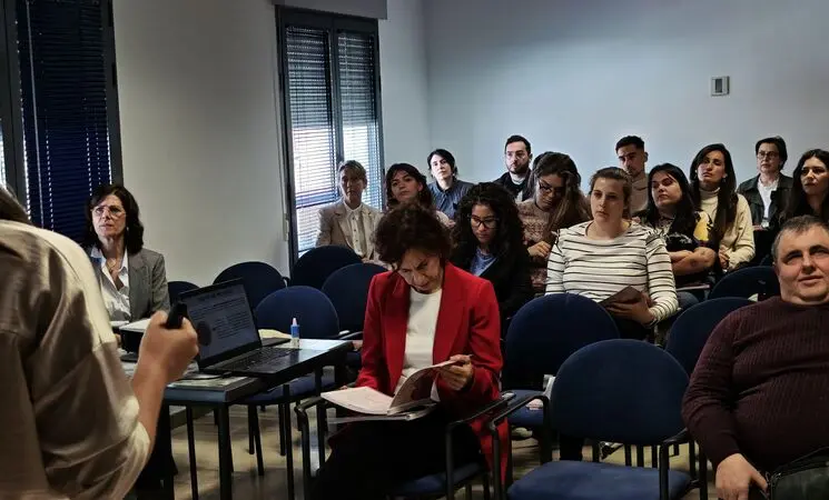 Imagen de una sala de formaci&oacute;n con varias personas sentadas. En primer plano, una persona habla de pie junto a una mesa con un ordenador port&aacute;til. Entre el p&uacute;blico, destaca una mujer con chaqueta roja leyendo un documento. El ambiente es formal y de atenci&oacute;n durante una jornada o reuni&oacute;n.