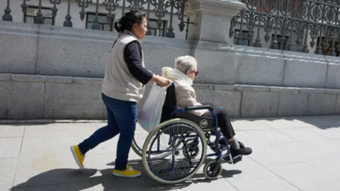 Imagen de una mujer mayor en silla de ruedas junto a su cuidadora paseando un 4 de junio de 2025 en Madrid