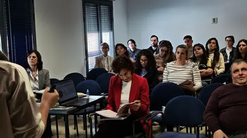 Imagen de una sala de formaci&oacute;n con varias personas sentadas. En primer plano, una persona habla de pie junto a una mesa con un ordenador port&aacute;til. Entre el p&uacute;blico, destaca una mujer con chaqueta roja leyendo un documento. El ambiente es formal y de atenci&oacute;n durante una jornada o reuni&oacute;n.